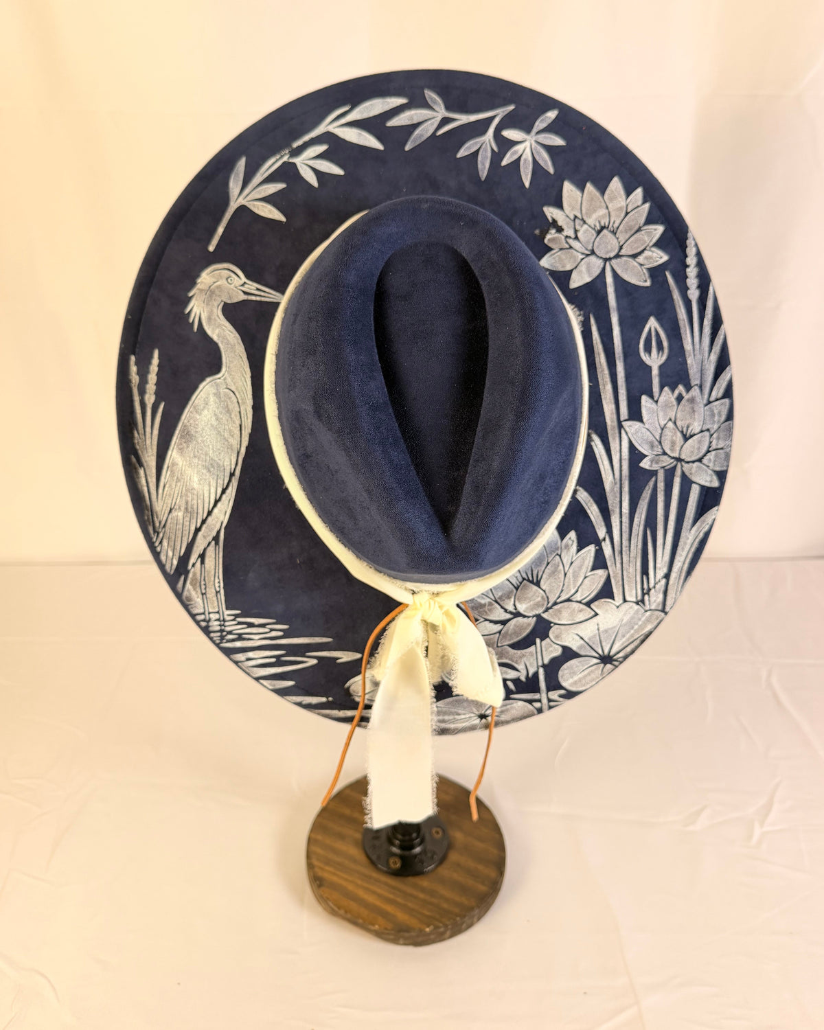 A wide-brimmed navy blue hat with white floral and bird illustrations, displayed on a wooden stand against a light beige backdrop.