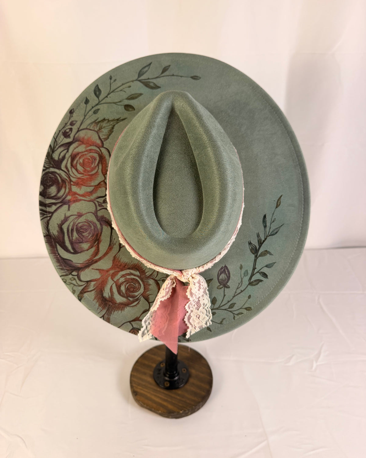 A wide-brimmed sage green cowboy hat with a floral rose pattern on the brim, displayed on a wooden stand. The hat features a pink ribbon and white lace trim at the base of the crown.