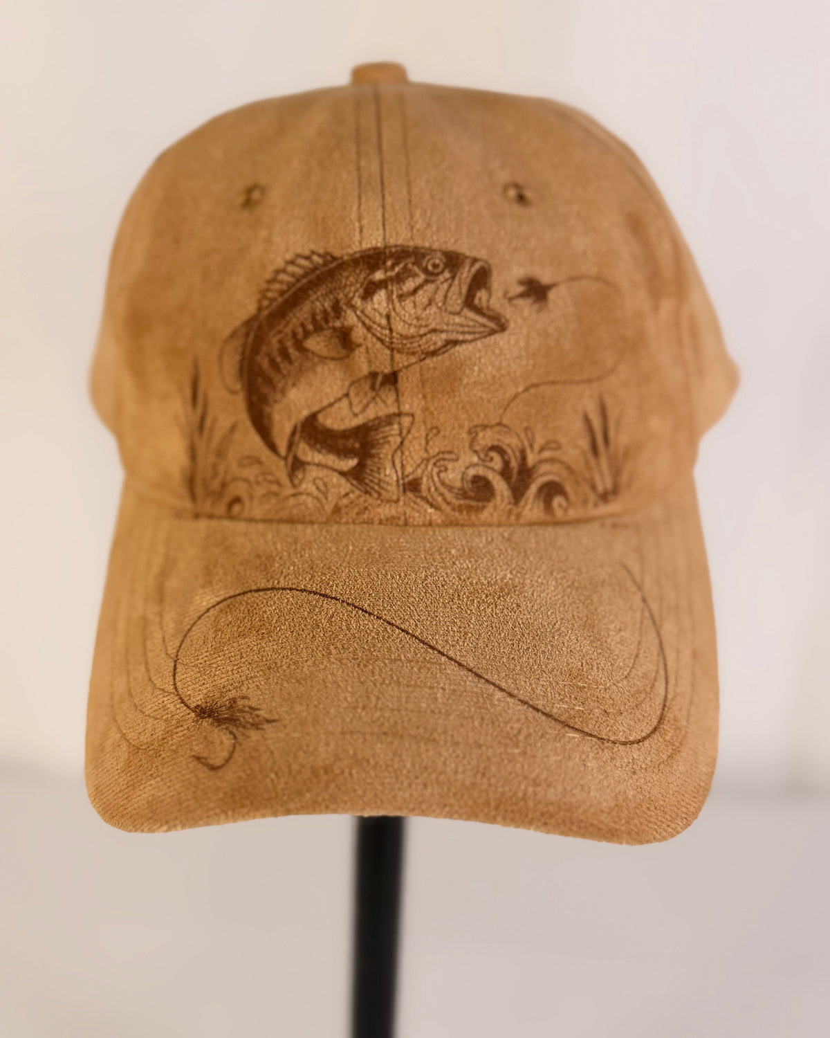 A tan baseball cap with a laser-engraved design of a largemouth bass fishing on a fly, displayed on a black stand against a white background.