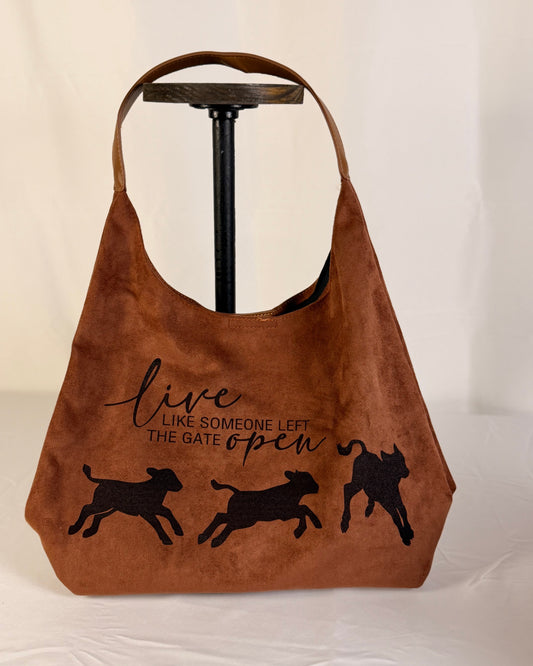 A brown suede hobo-style shoulder bag displayed on a black stand against a white fabric backdrop. The bag features black printed text reading 'live LIKE SOMEONE LEFT THE GATE open' above three black jumping calf silhouettes.