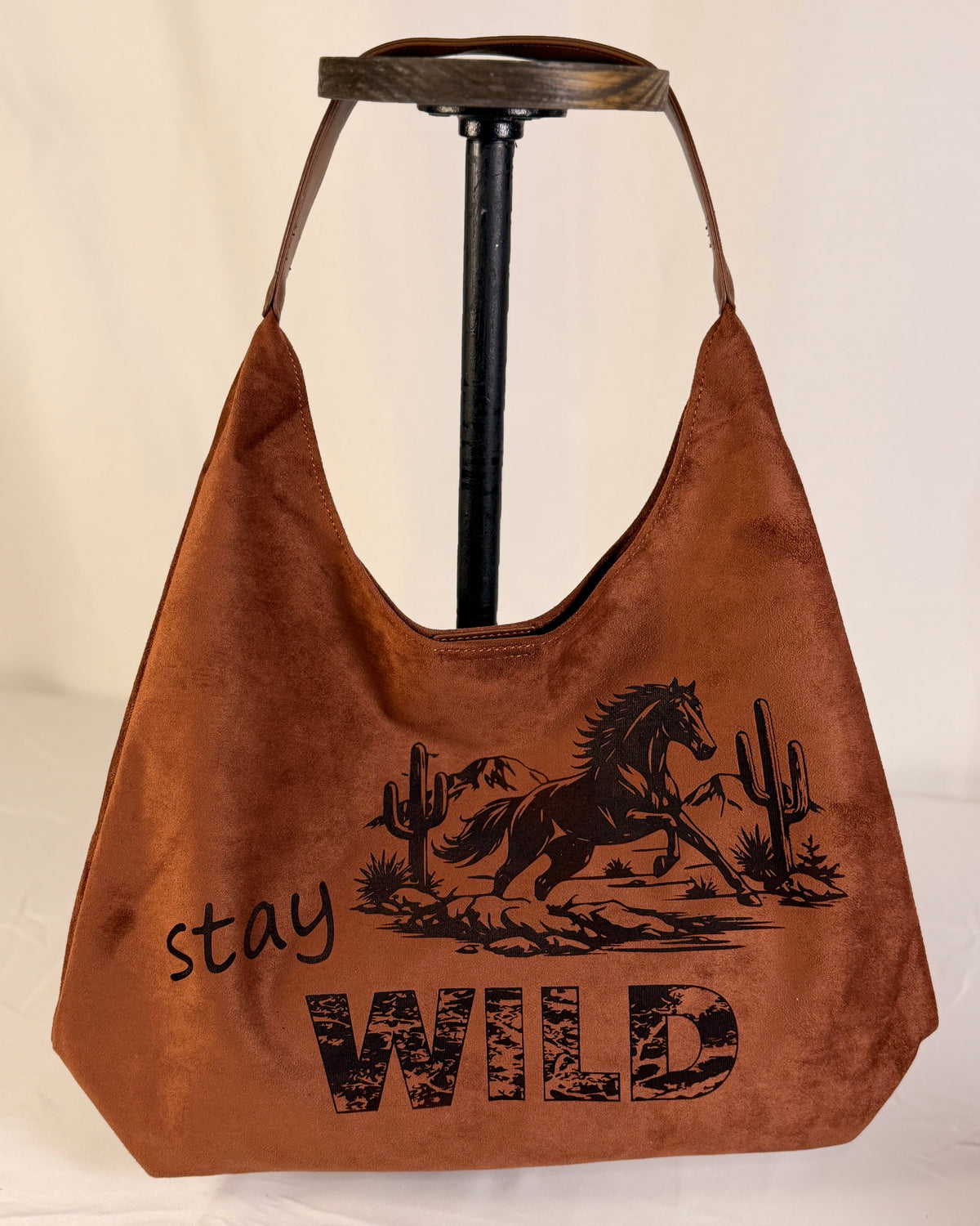 A brown suede hobo-style shoulder bag displayed on a black stand against a white fabric backdrop. The bag features a black graphic print of a prancing horse with cacti and the words 'stay WILD'.