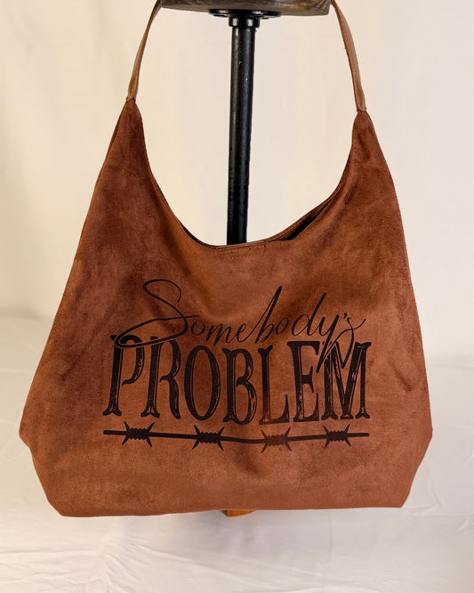 A brown suede hobo shoulder bag hanging on a black stand against a white fabric backdrop. The bag features the phrase 'Somebody's PROBLEM' printed in black across the front, with 'Somebody's' in cursive and 'PROBLEM' in a bold serif font above a barbed wire graphic.