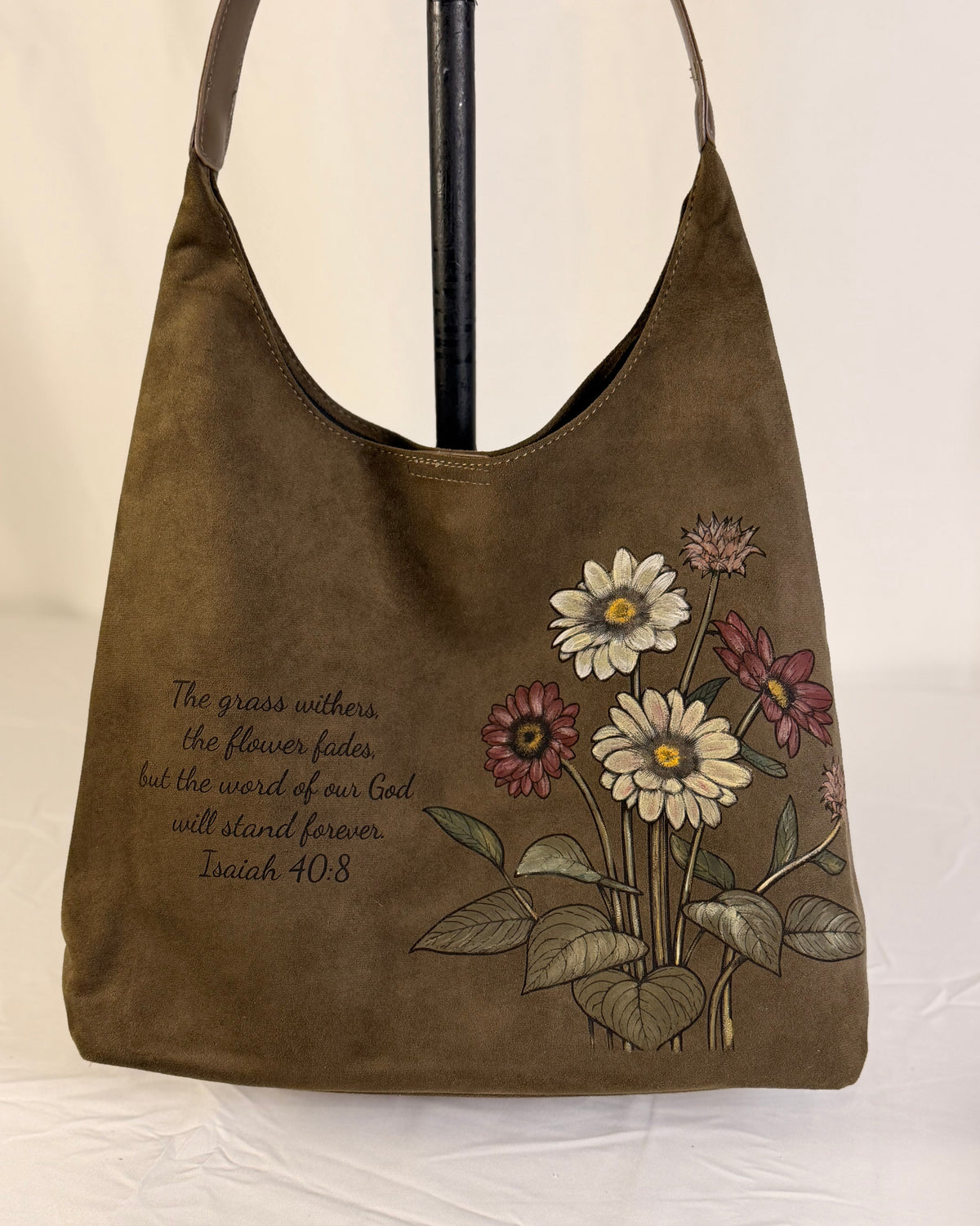 An olive brown suede hobo shoulder bag featuring a floral design with white, pink, and green flowers, a Bible verse printed on the left, and a brown shoulder strap.