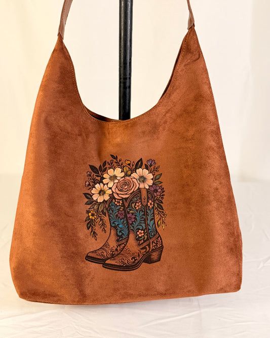 A brown suede hobo shoulder bag with a floral cowboy boot graphic, displayed on a black stand against a white fabric backdrop.
