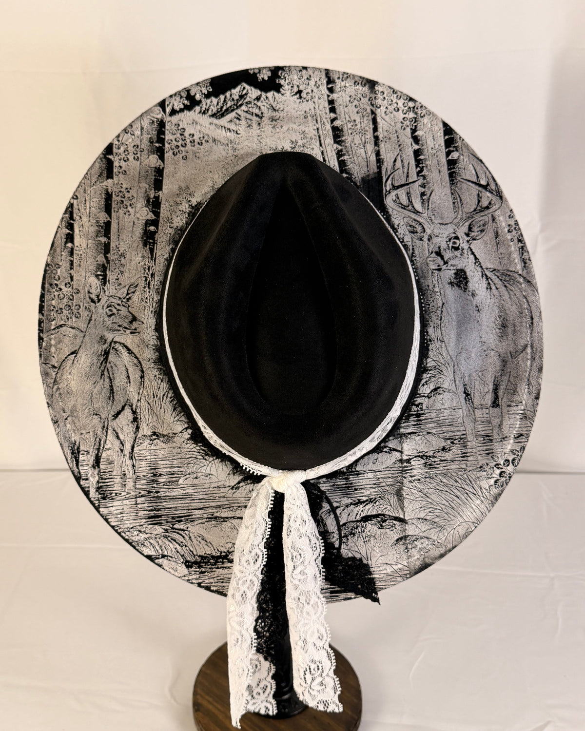 A black wide-brim hat with a black and white forest and deer print, featuring a white lace ribbon tie, displayed on a wooden stand against a white background.