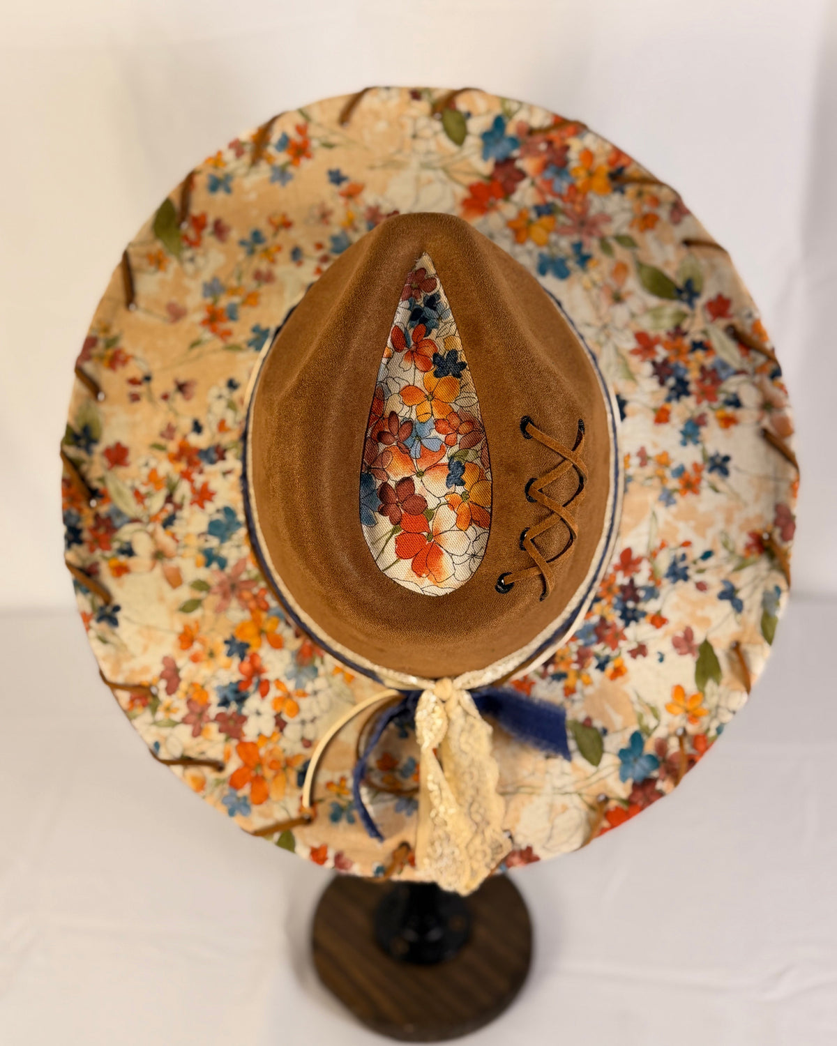 A handmade wide-brimmed hat with a brown felt crown and a floral patterned fabric brim, displayed on a wooden stand against a white background.